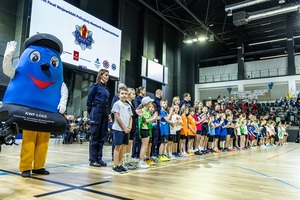 Policjanci z zawodnikami i maskotką Komisarzem Błyskiem stoją wzdłuż linii na hali sportowej.
