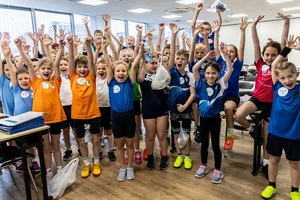 Dzieci na sali z ławkami machają do zdjęcia.