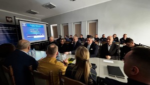 Przedstawiciele samorządów siedzą przy biurkach, z tyłu siedzą policjanci, po lewej stronie wyświetlana jest prezentacja.