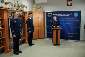 Komendant przemawia za mównicą, po jego lewej stronie stoją zastępcy.